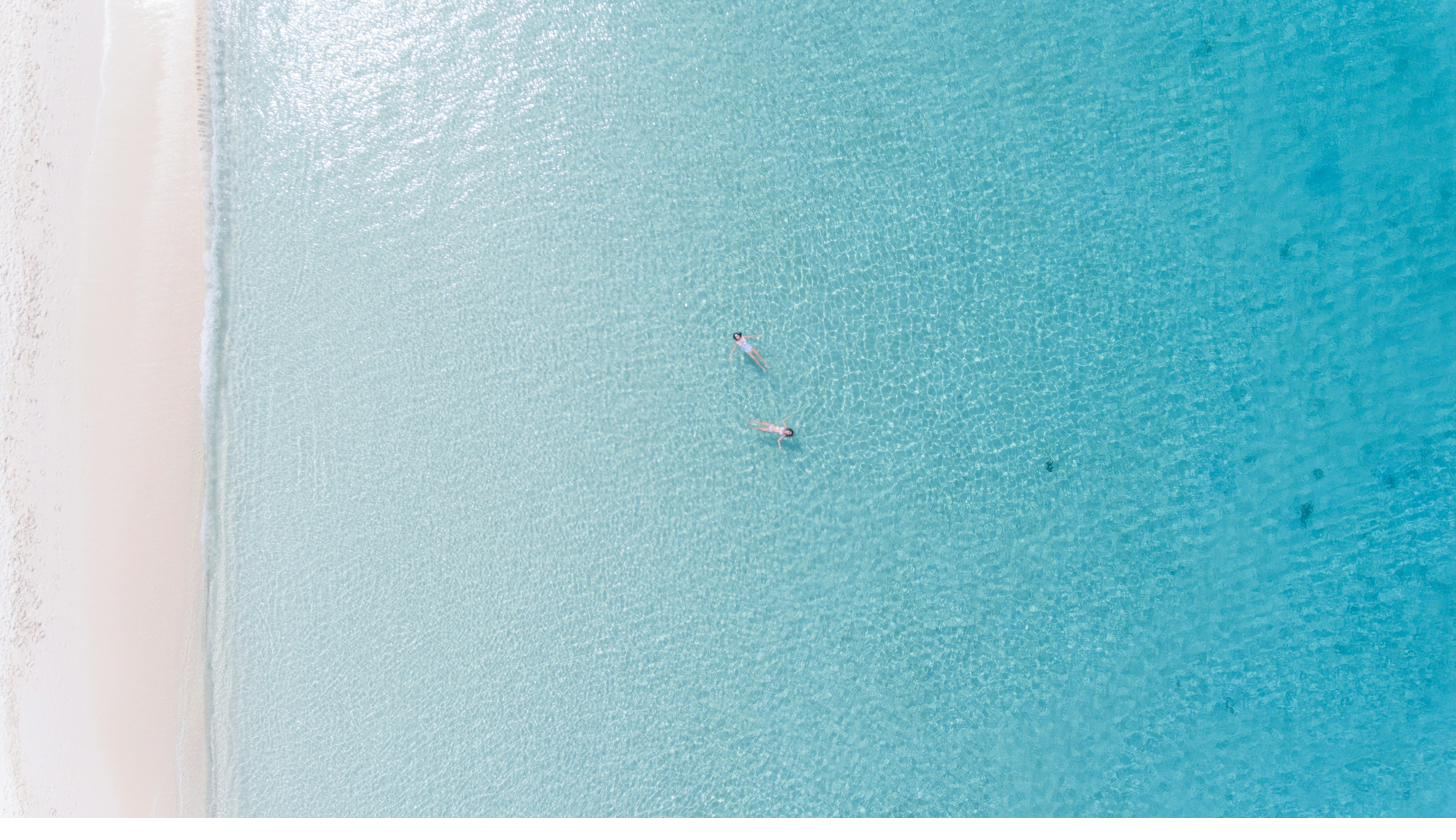 Two people floating in clear turquoise water off a Cayman beach, seen from above